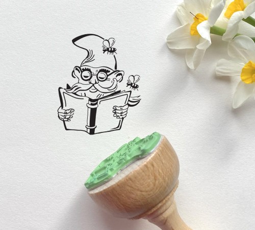 Pieczątka drewniana z czytającym krasnalem z Wrocławia. Krasnal przedstawiony jest w okularach i czapce, w dłoniach trzyma książkę, a wokół niego  latają pszczółki.